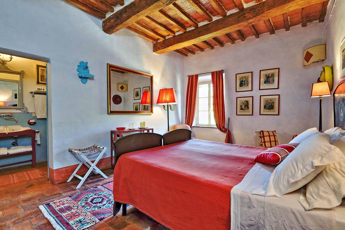A bedroom with a large bed, a desk, a chair, a small table, and a window with curtains.