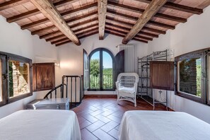 A room with a high ceiling, wooden beams, a bed, a chair, a radiator, and a window with a view.