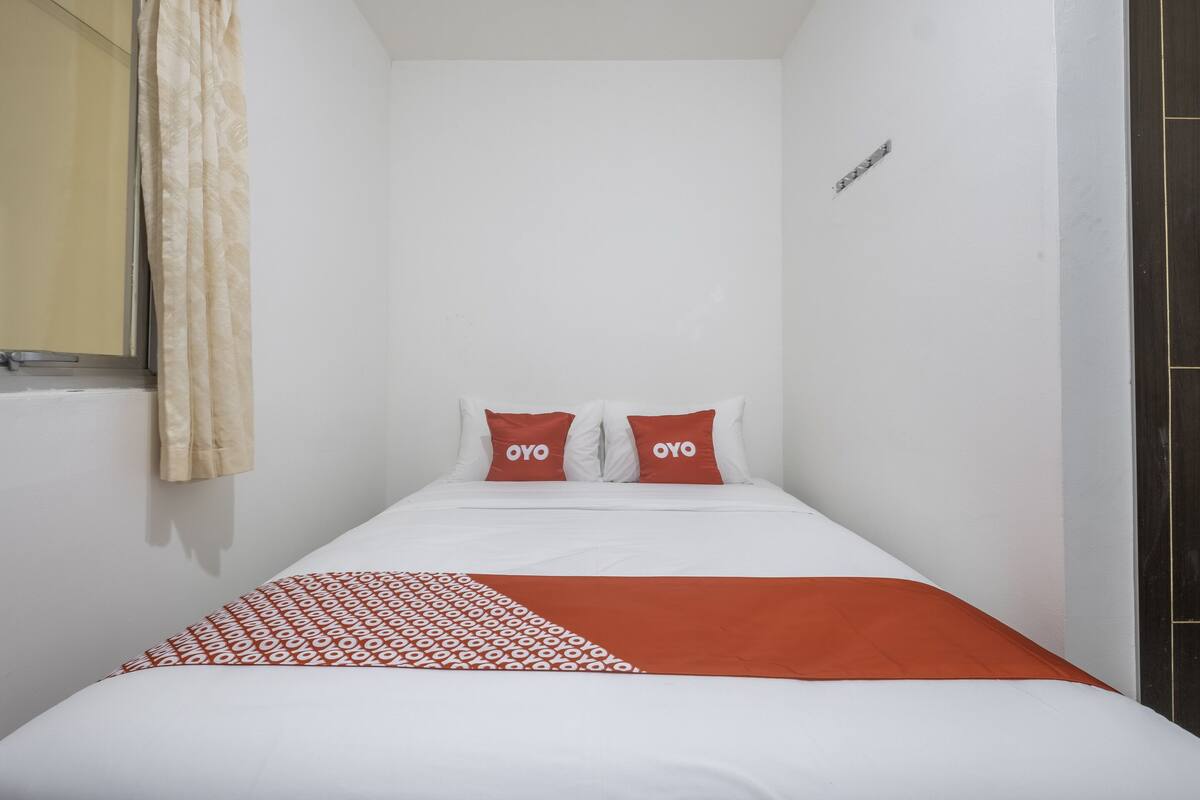 A small, single-bed room with a white and orange bedspread, two matching pillows, and a beige curtain.