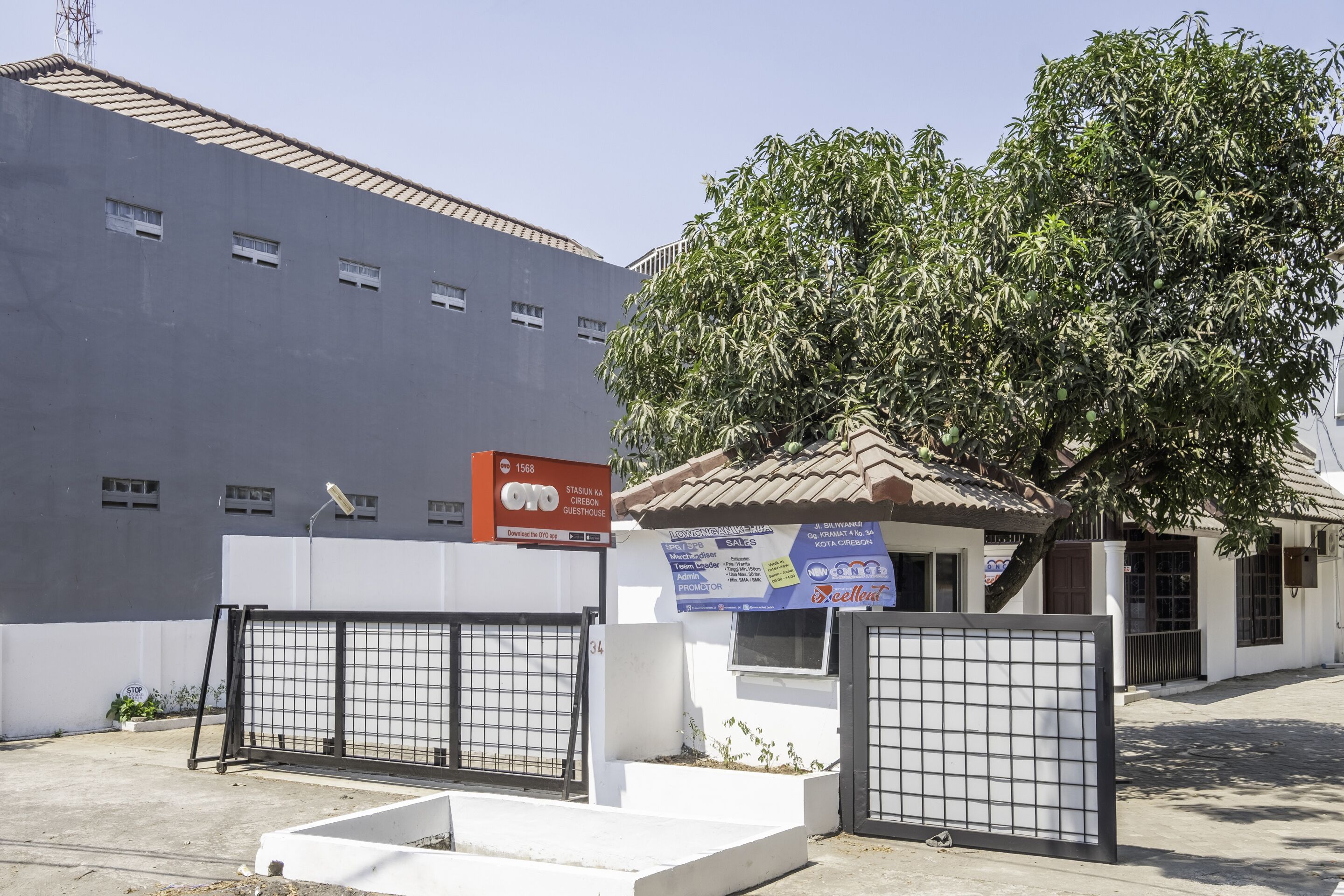 A building with a grey facade, a tiled roof, and a tree in front. There is a sign on the building and a gated entrance.