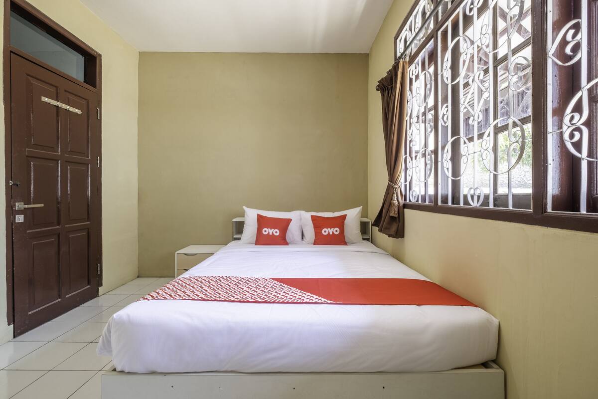 A neatly made bed with white and red bedding, a brown door, and a window with decorative iron bars.