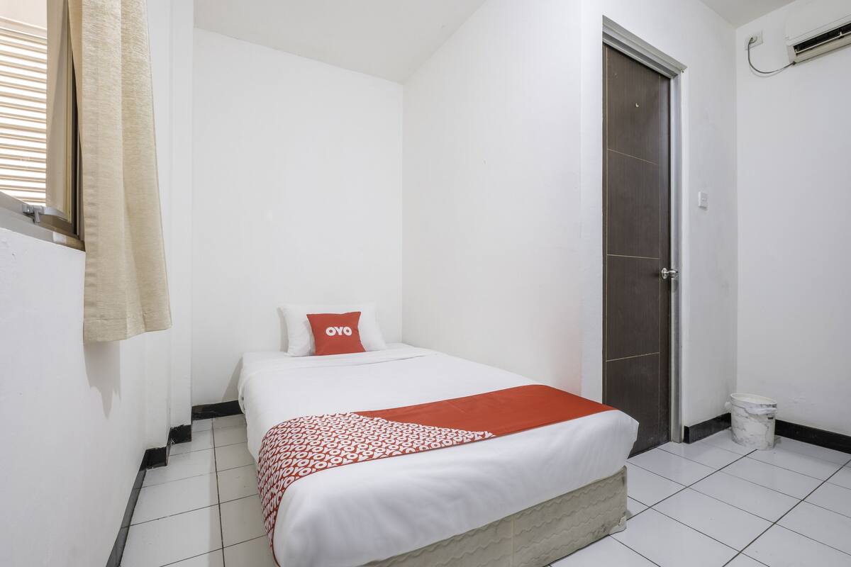 A hotel room with a bed, a door, a window with blinds, and a trash bin.