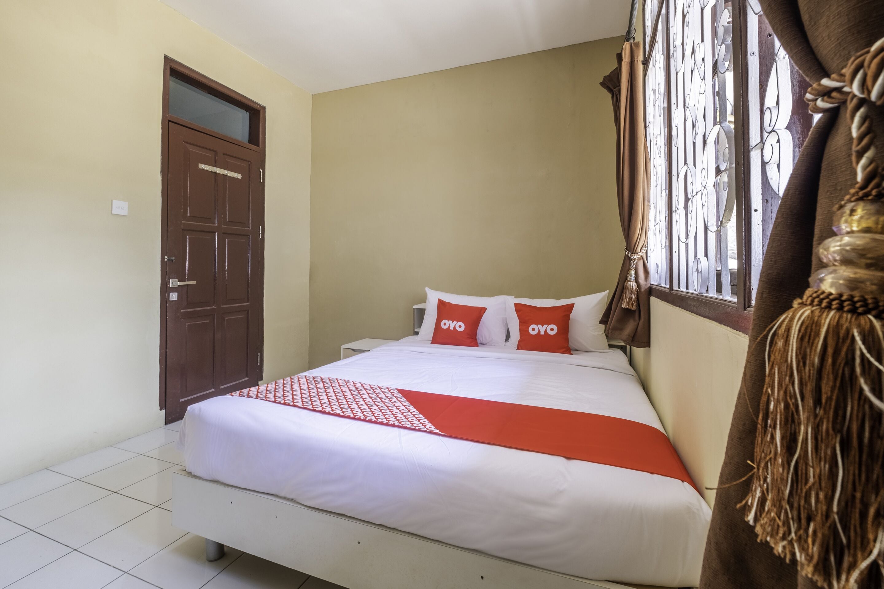 Une chambre avec un lit, deux oreillers portant la marque « OYO », une fenêtre avec des barreaux en métal, un rideau marron et une porte.