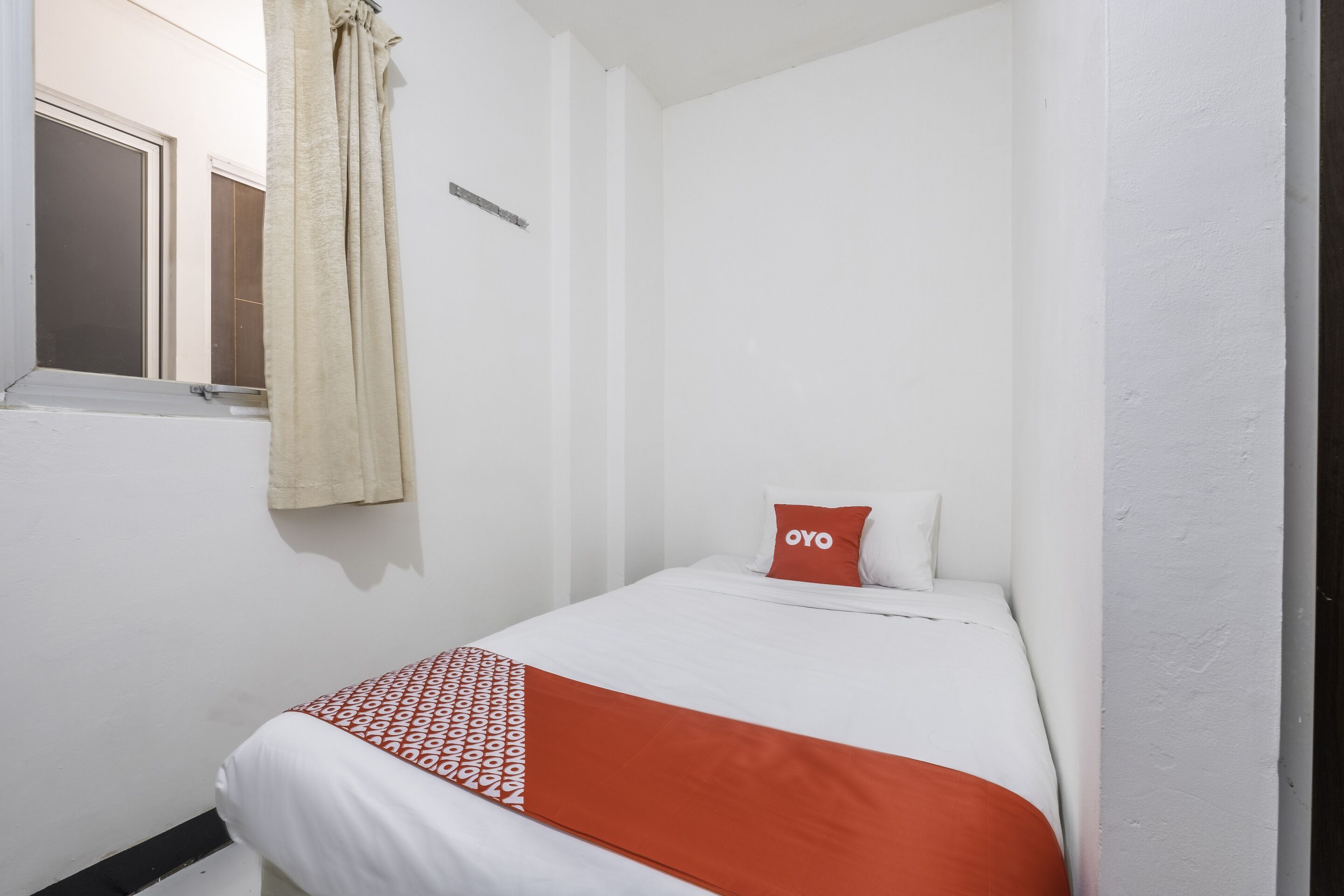 A small, single-bed room with white walls, a window with a curtain, and a bed with a red and white patterned blanket.