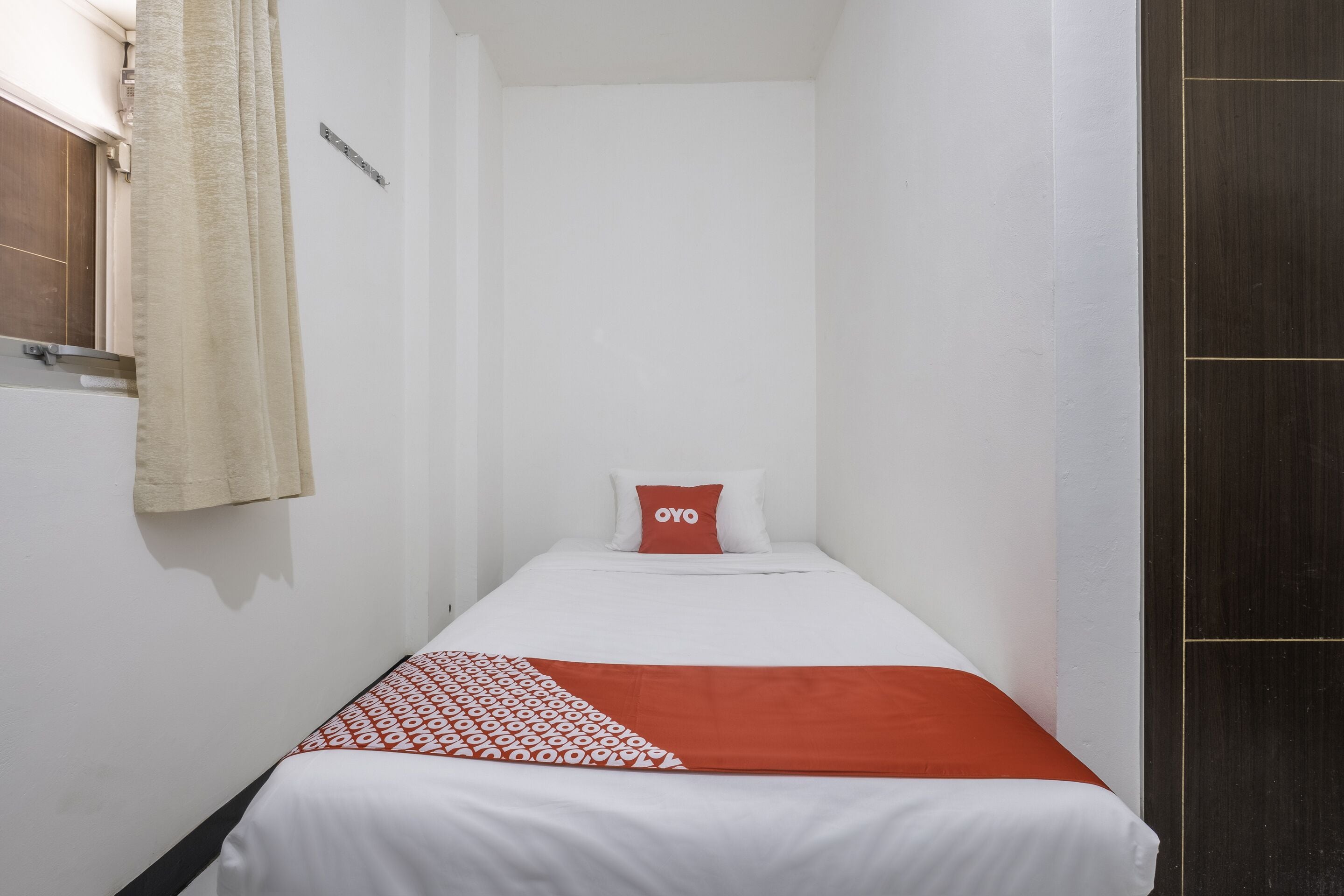 Una habitación pequeña con cama individual, una colcha blanca, un almohadón rojo y una manta estampada.