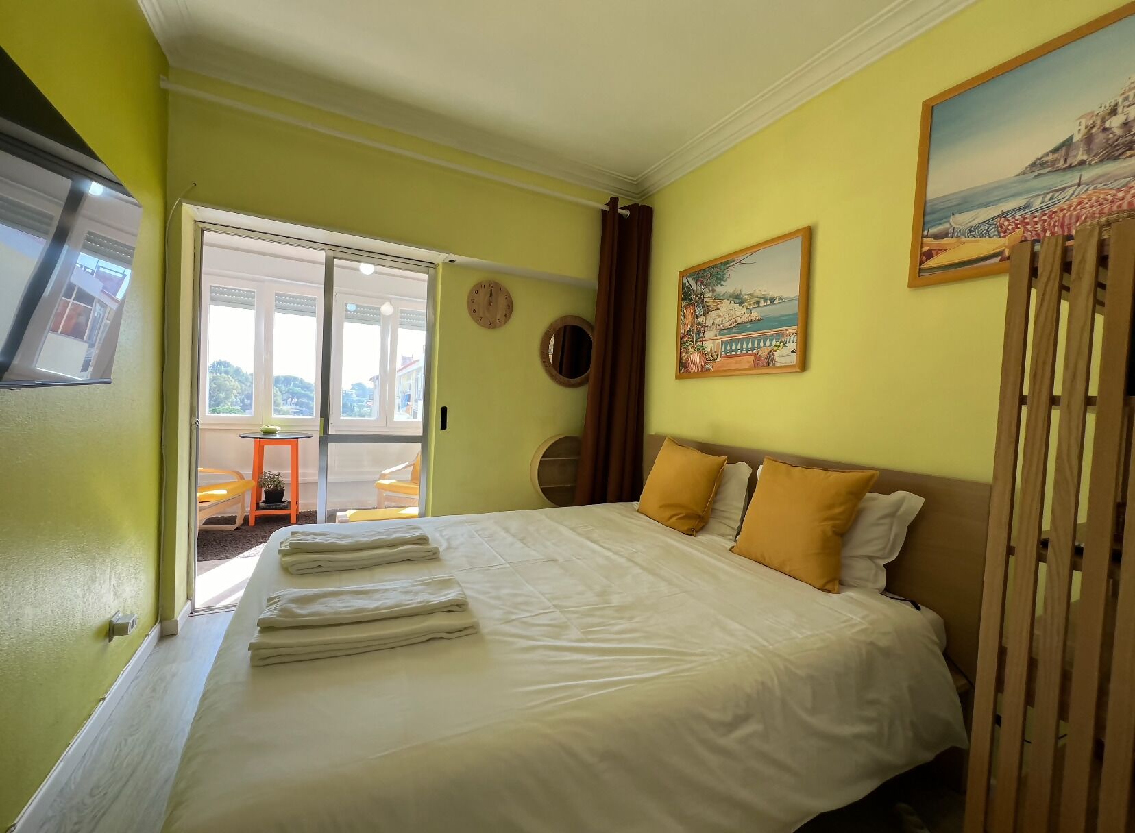 A hotel room with a bed, a painting, a clock, and a view of the outside.