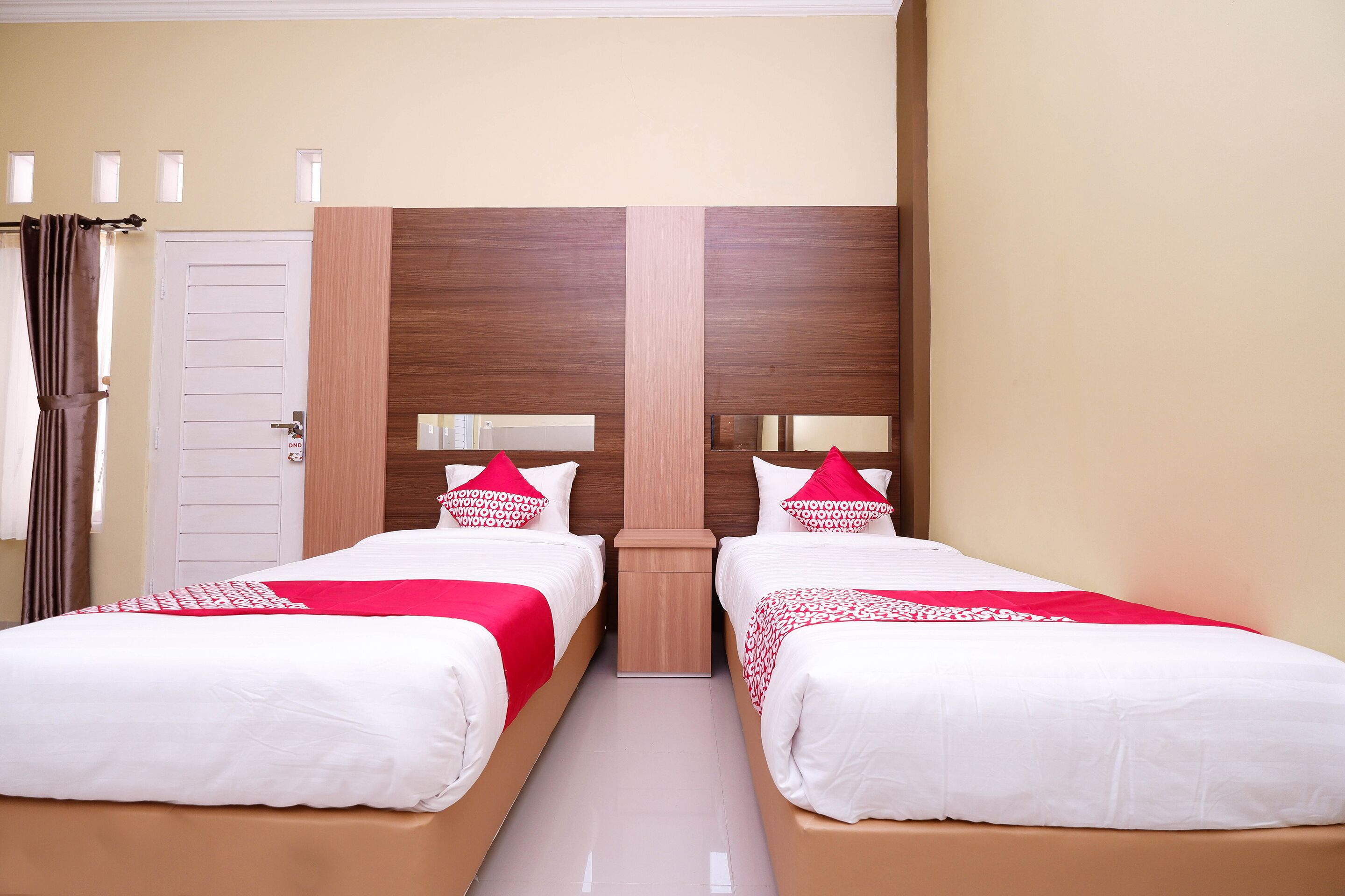 A hotel room with two single beds, a wooden headboard, a white door, and a small bedside table.