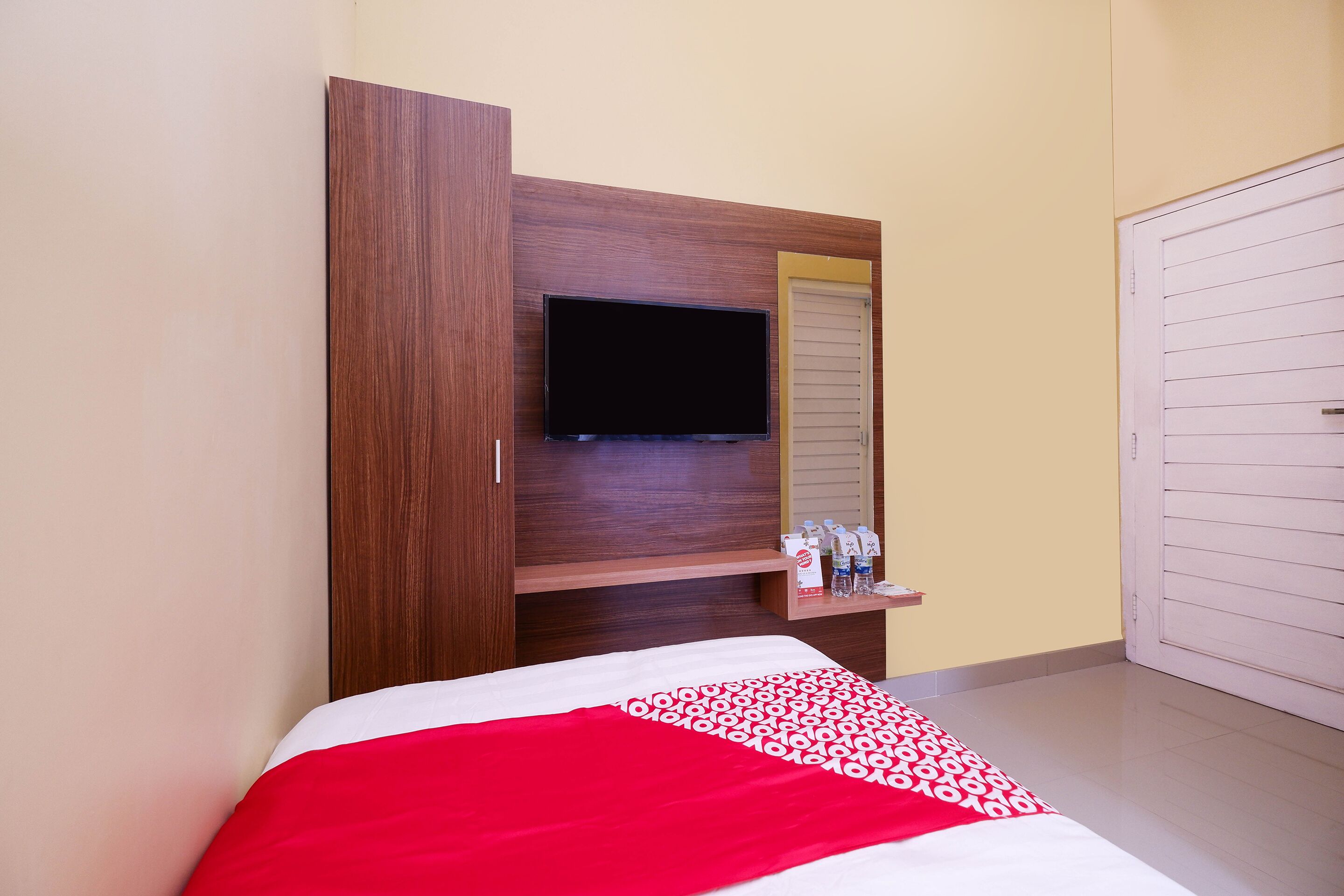 A hotel room with a bed, a TV mounted on a wooden panel, a shelf with toiletries, and a door with a sliding shutter.