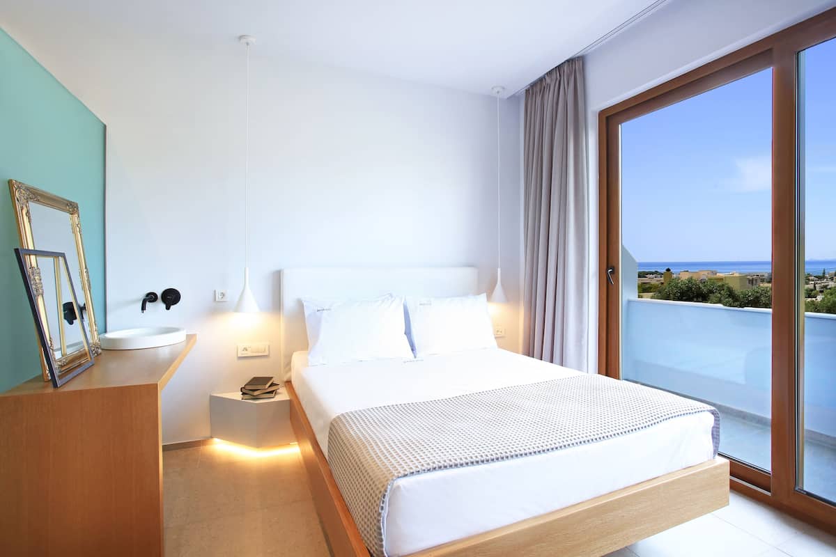 A modern hotel room with a large bed, a bedside table, a nightstand, a bathroom mirror, and a view of the sea through a sliding glass door.