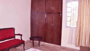 A room with a wooden wardrobe, a red upholstered bench, a small round table, and a window with curtains. - Hotel Residence du Bonheur (Abidjan)