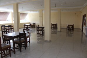 A spacious dining area with multiple wooden tables and chairs, a tiled floor, and large windows with curtains.