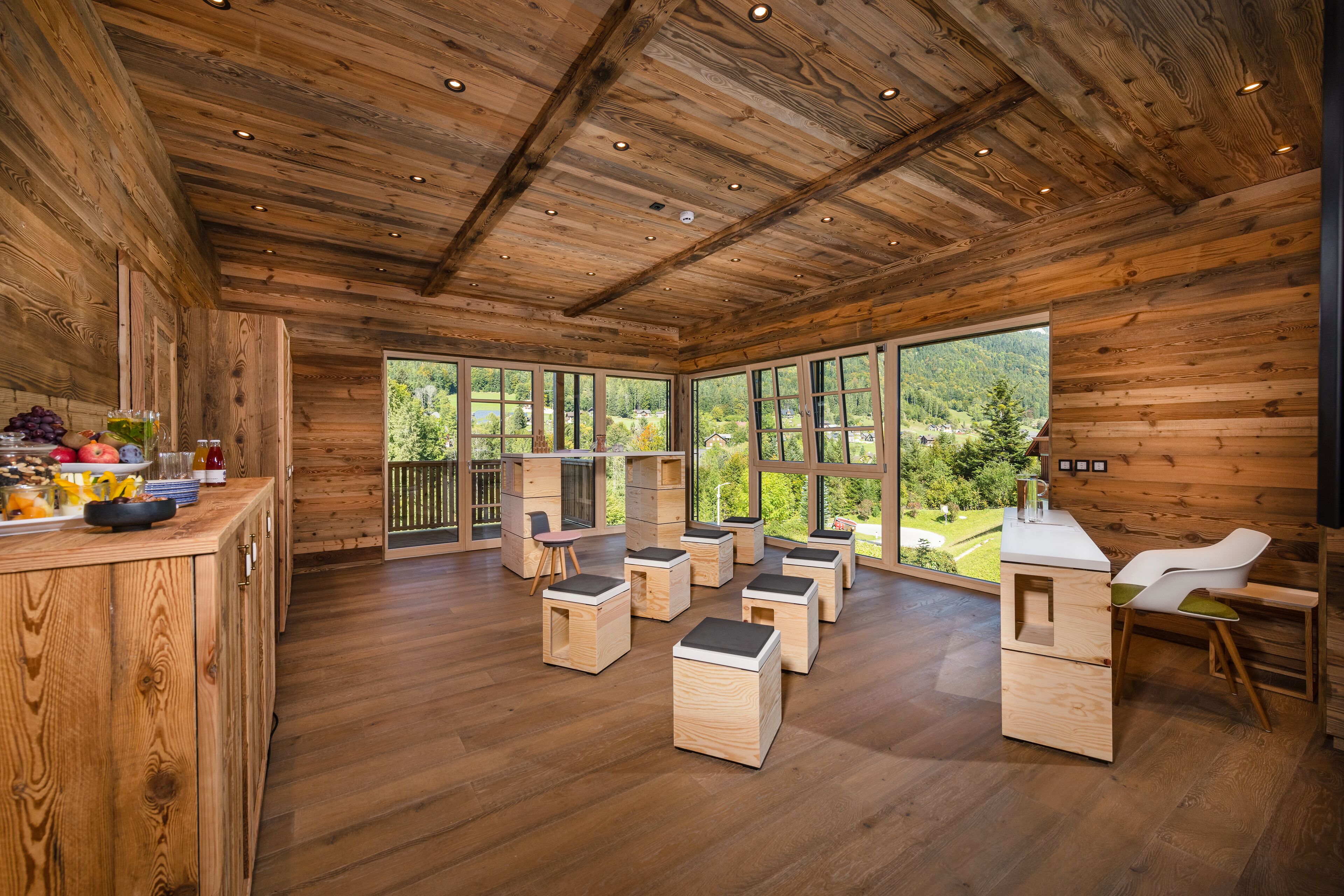 A wooden cabin interior with a kitchen area, dining space, and large windows offering a view of the outdoors.