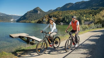 Dos ciclistas en un camino pavimentado junto a un lago, con montañas al fondo.
