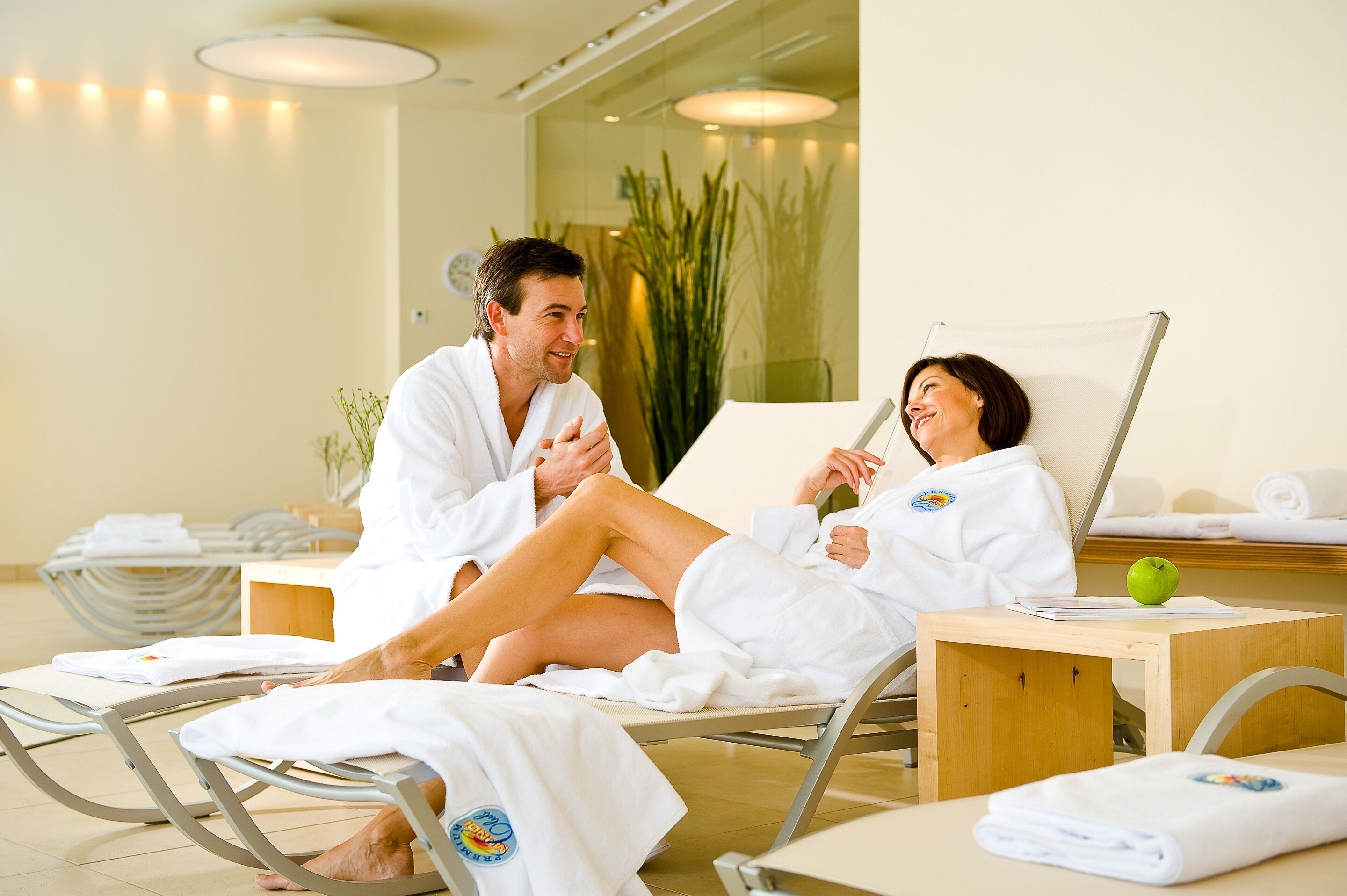 A spa setting with two people in white robes seated on reclining chairs, a wooden table with a green apple, and a large plant in the background.