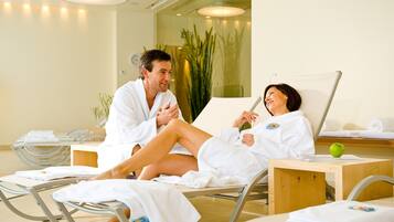 A spa setting with two people in white robes seated on reclining chairs, a wooden table with a green apple, and a large plant in the background.