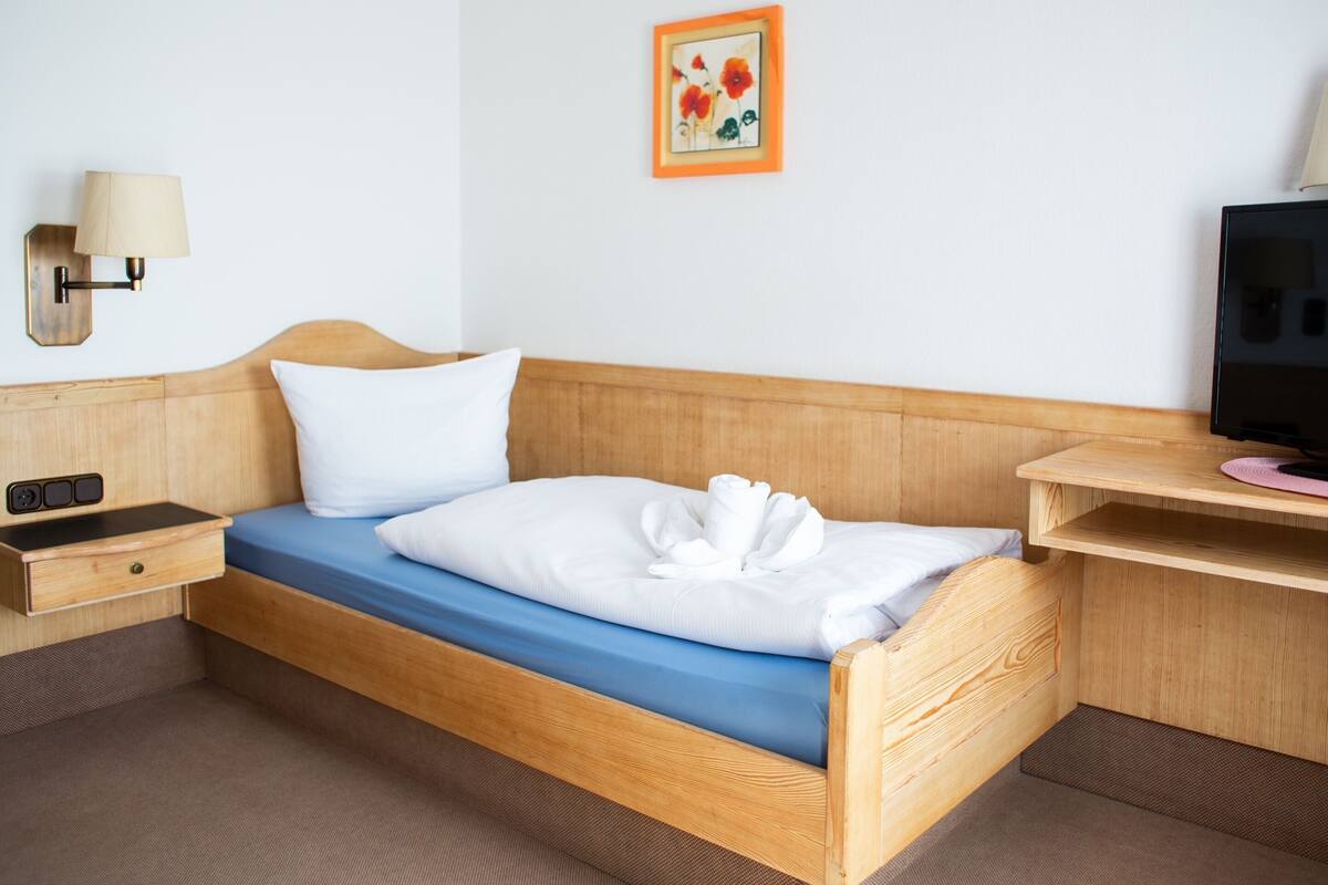 En seng med træstel, blå madras, hvide puder og et håndklæde. Der er et trænatbord med en lampe, et fjernsyn på et træstel og et indrammet billede på væggen.