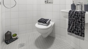A modern bathroom with white tiles, a toilet, a shower, and a sink with a towel. - Yugo Explore - Broadstone Hall (Dublin)
