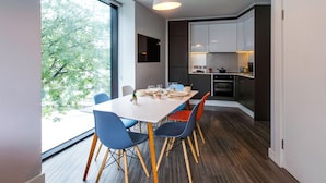 A modern kitchen with white cabinets, a dining area with a white table and blue chairs, and a large window with a view of trees. - Yugo Explore - Broadstone Hall (Dublin)
