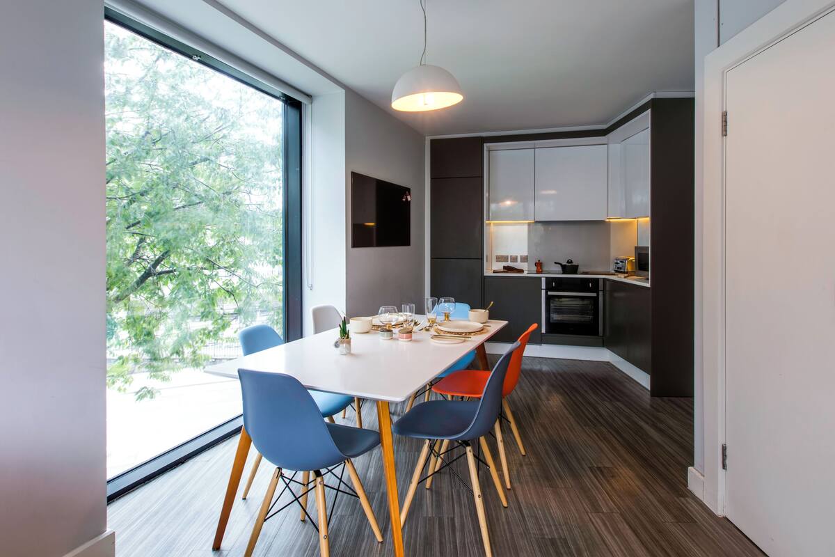 A modern kitchen with white cabinets, a dining area with a white table and blue chairs, and a large window with a view of trees.