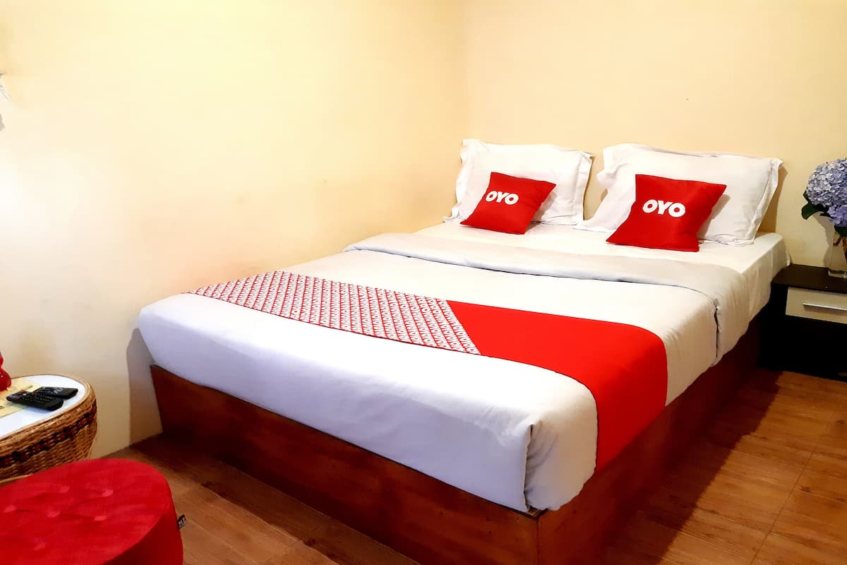 A neatly made bed with white and red bedding, two red pillows with the word "OYO" on them, a red throw blanket, and a small wicker side table with a remote control.
