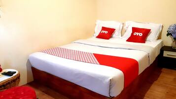 Una cama bien hecha con ropa blanca y roja, dos almohadas rojas con la palabra "OYO" estampada, un plaid rojo y una pequeña mesita de mimbre con un control remoto.