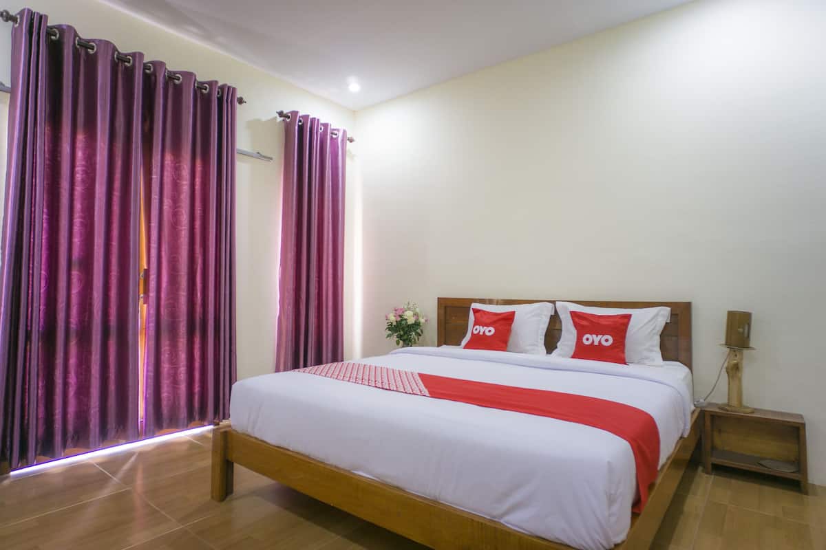 A hotel room with a large bed, two pillows with 'OYO' branding, a nightstand with a lamp, and purple curtains.