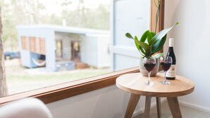 A round wooden table with two wine glasses and a bottle of wine, a potted plant, and a window with a view of a building outside. - Tuckers Lane Boutique Accommodation (North Rothbury)