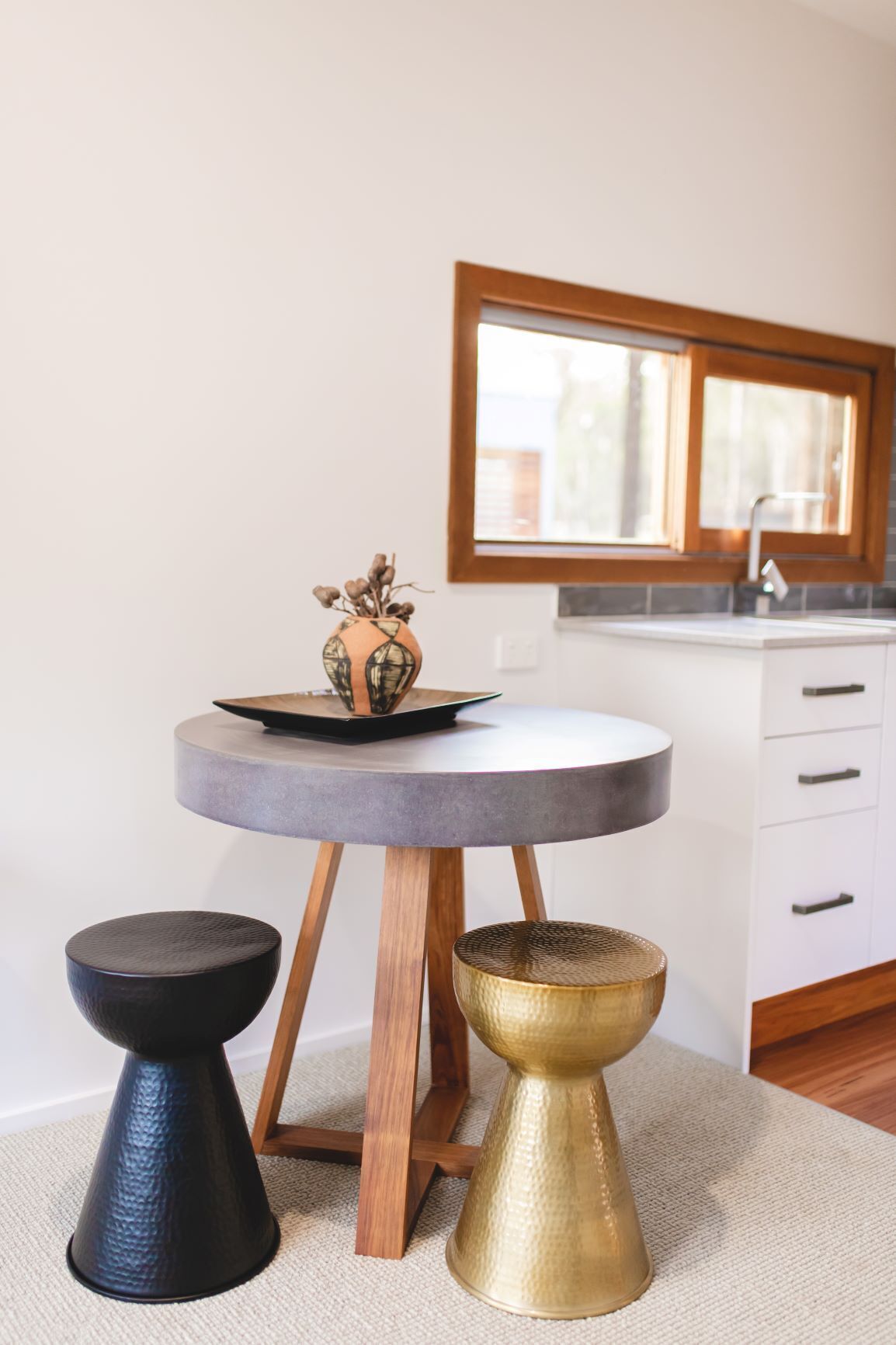 A round table with a vase and a black object on top, flanked by two stools, in a room with a window and a white cabinet.