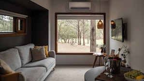 A modern living room with a grey sofa, a round coffee table, and a view of a forest through a large window. - Tuckers Lane Boutique Accommodation (North Rothbury)