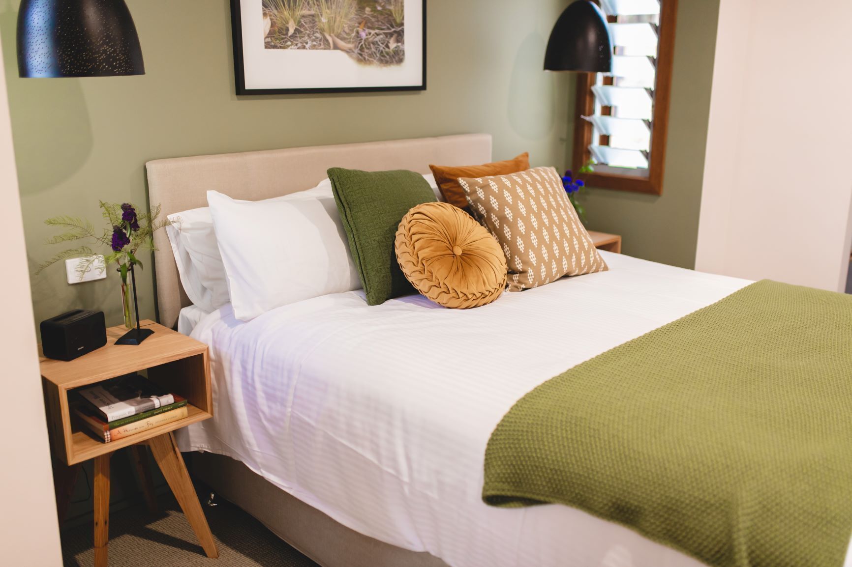 A neatly made bed with a white comforter, multiple pillows, and a green throw blanket. There is a wooden nightstand with a lamp and a small plant beside the bed.
