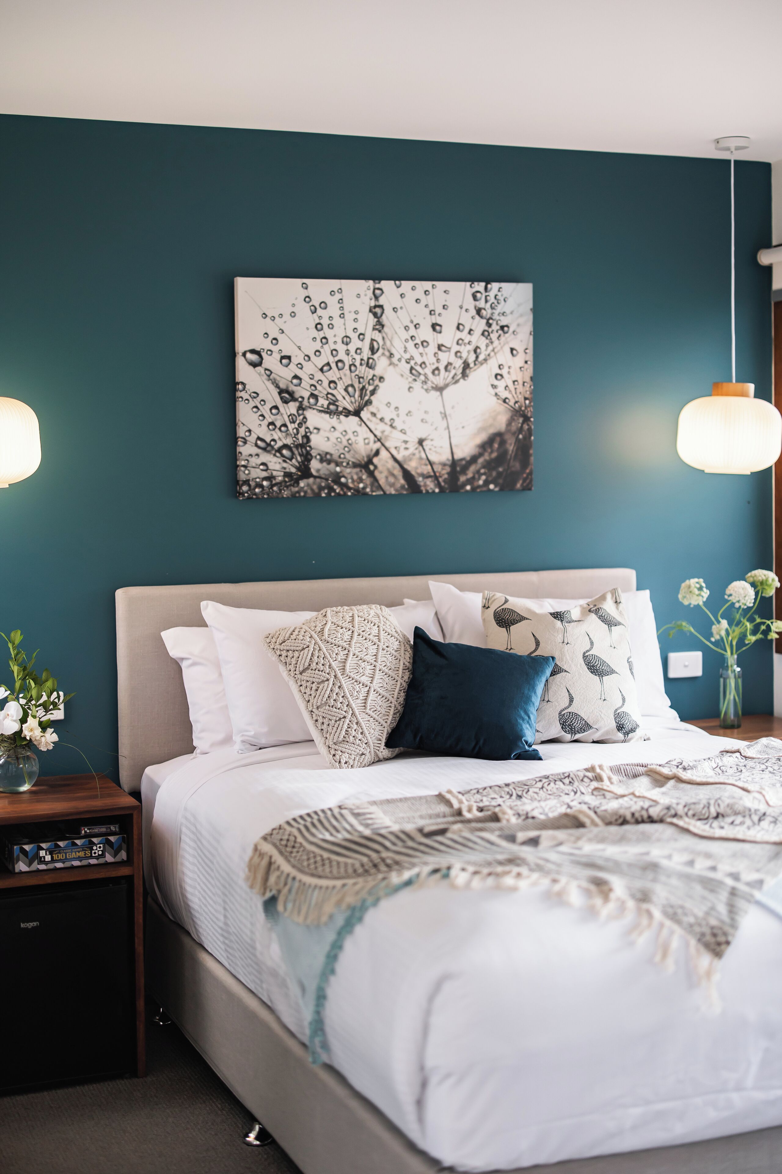 Ein ordentlich bezogenes Bett mit einer gemusterten Tagesdecke, ein an der Wand befestigtes Kunstwerk und ein Nachttisch mit Lampe.
