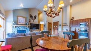 A modern living room with a stone fireplace, a wooden dining table, and a kitchen area with wooden cabinets and a microwave.