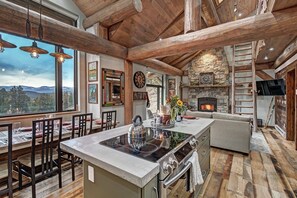 A spacious kitchen with a central island, a stone fireplace, and a large window overlooking a mountainous landscape. - Skyrun 2 Bedroom Cabin by RedAwning (Golden)