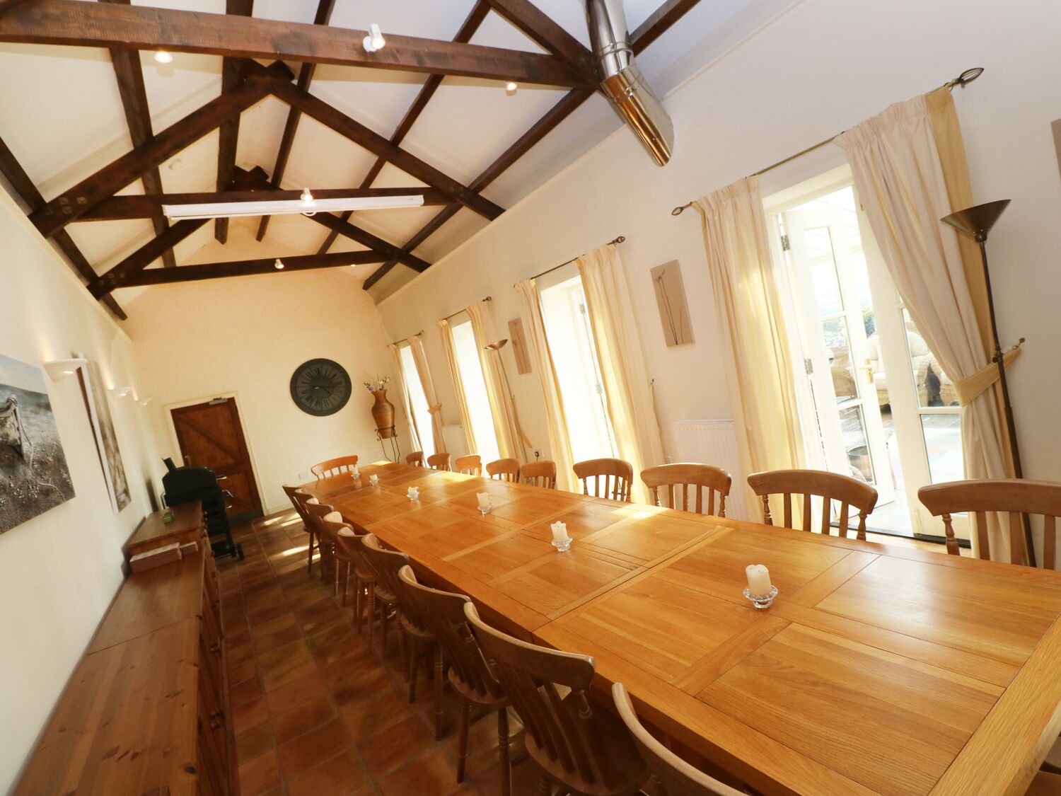 A large wooden dining table with chairs, a wooden ceiling with exposed beams, and large windows with curtains.