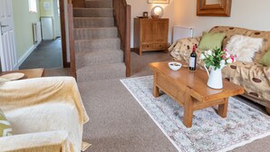 Un salon chaleureux avec un escalier en bois, un tapis à motifs, une table basse en bois ornée d’un vase de fleurs et un canapé avec des coussins à motifs.