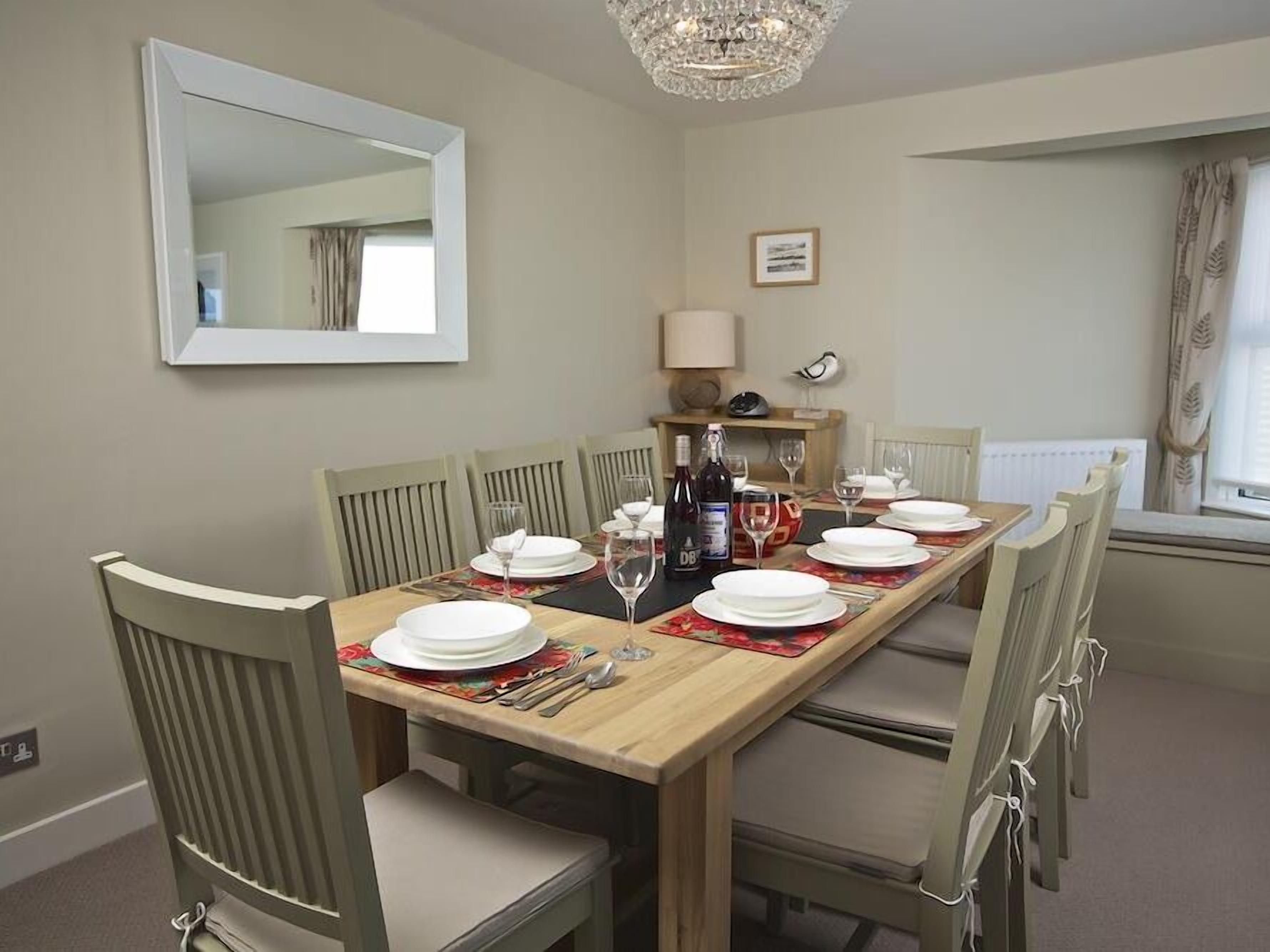 A dining room with a wooden table set for six, surrounded by six chairs. A chandelier hangs from the ceiling, and a mirror is mounted on the wall.