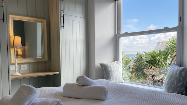 A hotel room with a large bed, a mirror, a window with a view of palm trees and a building, and a bedside table with a lamp. - Porth House with south facing courtyard garden! (St Ives)