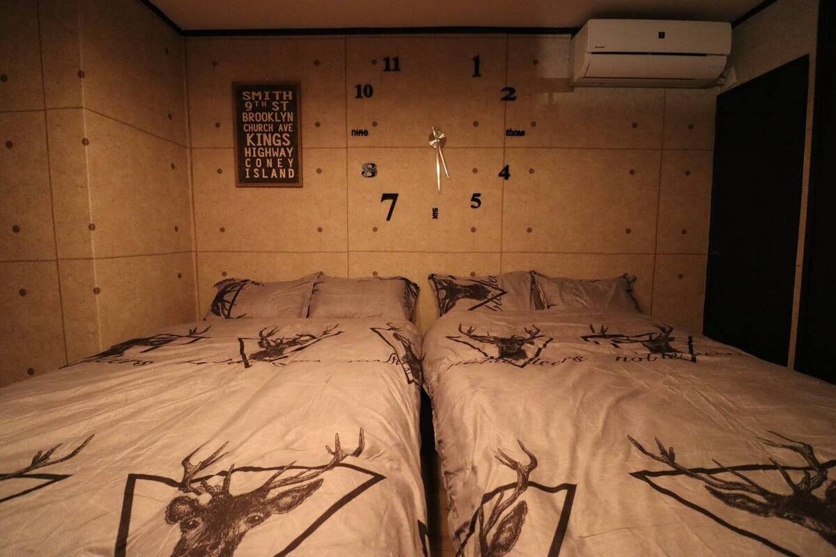 A room with two beds, each with a deer print bedspread. A wall clock is centered above the beds, and a sign above the clock lists addresses.