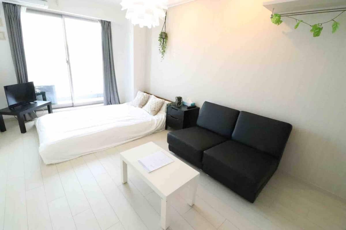 Una sala de estar moderna con una cama, un sofá negro, una mesa de centro blanca y un televisor sobre un mueble.