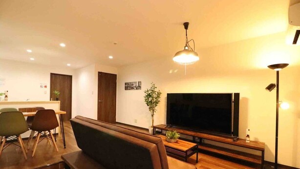 A modern living room with a large flat-screen TV, a wooden coffee table, a brown sofa, and a dining area with chairs and a small table. - Sky Terrace Kujyo (Osaka)