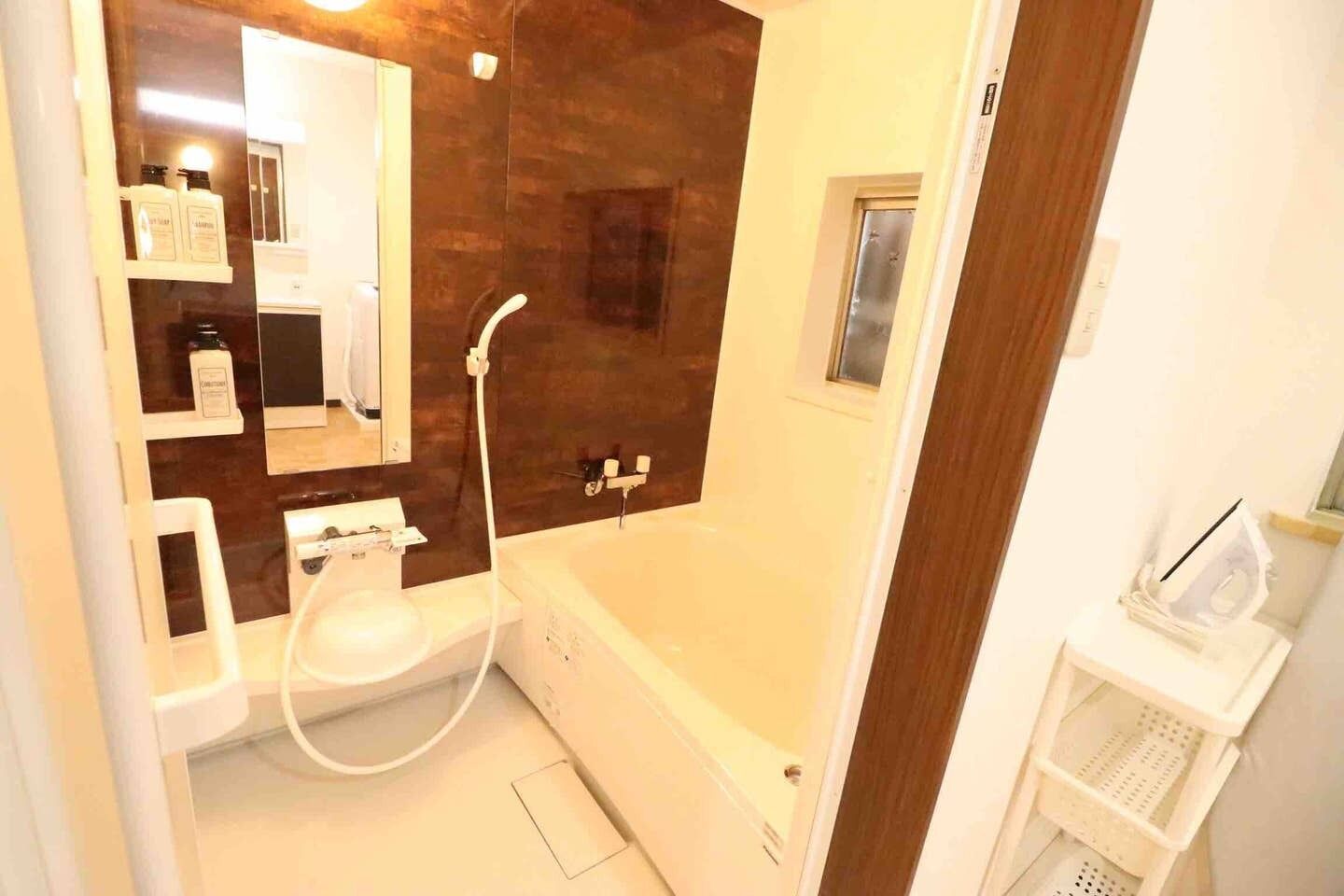 Une salle de bain avec une baignoire, un lavabo et un miroir. On y trouve une étagère avec des articles de toilette et une petite fenêtre.