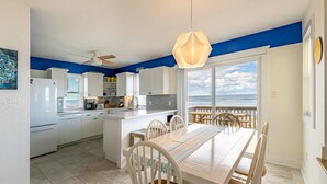 A modern kitchen with white cabinets, a central island, and a dining area with a view of the ocean. - Sun Trippin' | Oceanfront Escape in South Nags Head with Private Beach Access & Panoramic Views (South Nags Head)