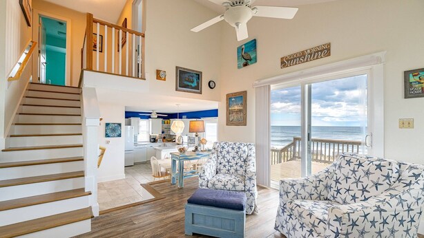 A spacious living area with a view of the ocean, a kitchen in the background, and a staircase leading to an upper level. - Sun Trippin' | Oceanfront Escape in South Nags Head with Private Beach Access & Panoramic Views (South Nags Head)