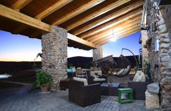 Un patio al aire libre con pilares de piedra, vigas de madera, mobiliario de mimbre y una noria grande.