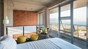 A modern bedroom with a large bed, a view of the city, a brick wall, and a balcony with a railing. - The Bond Square (Durban)