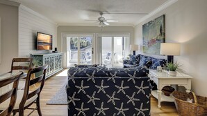 A living room with a ceiling fan, a TV mounted on the wall, a sofa with starfish patterns, a dining area with wooden chairs, and a view through the glass doors.