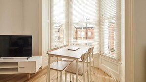 A corner room with a TV, a table, and chairs, large windows with blinds, and a view of buildings outside. - Host & Stay | The Normanby (Whitby)