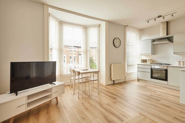 Une cuisine moderne équipée d’une cuisinière, d’un four et de meubles blancs. On y trouve une table à manger avec des chaises, ainsi qu’un téléviseur à écran plat posé sur un meuble blanc.