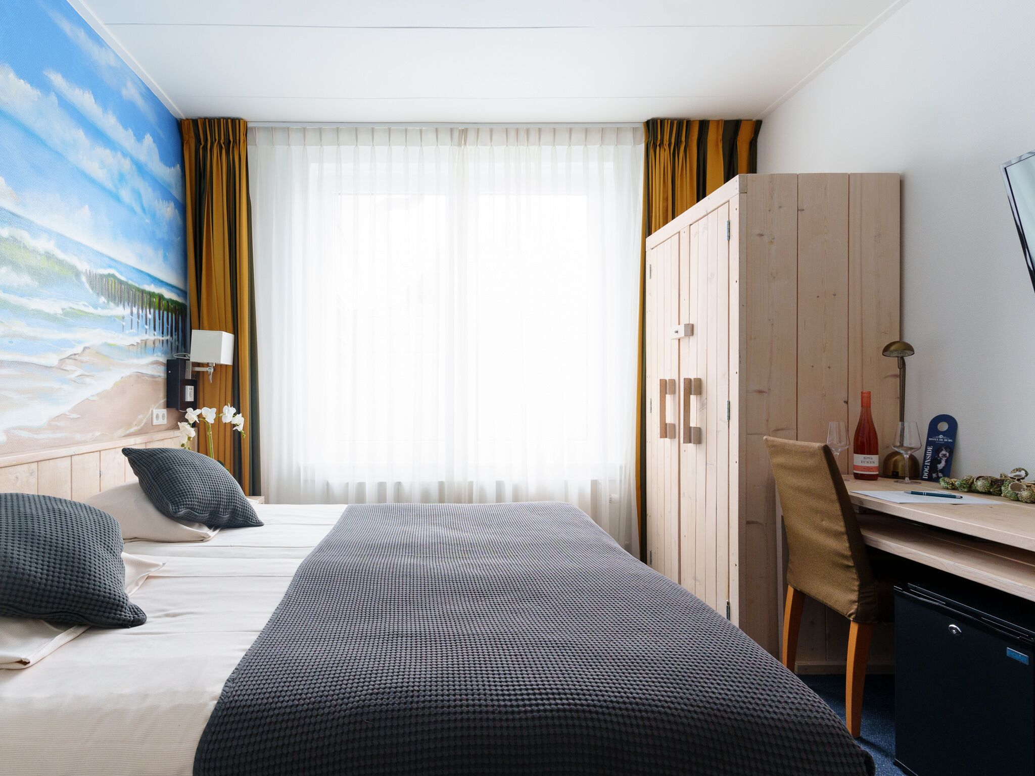 Une chambre d’hôtel avec un lit, un bureau, une armoire et un tableau représentant une scène de plage.