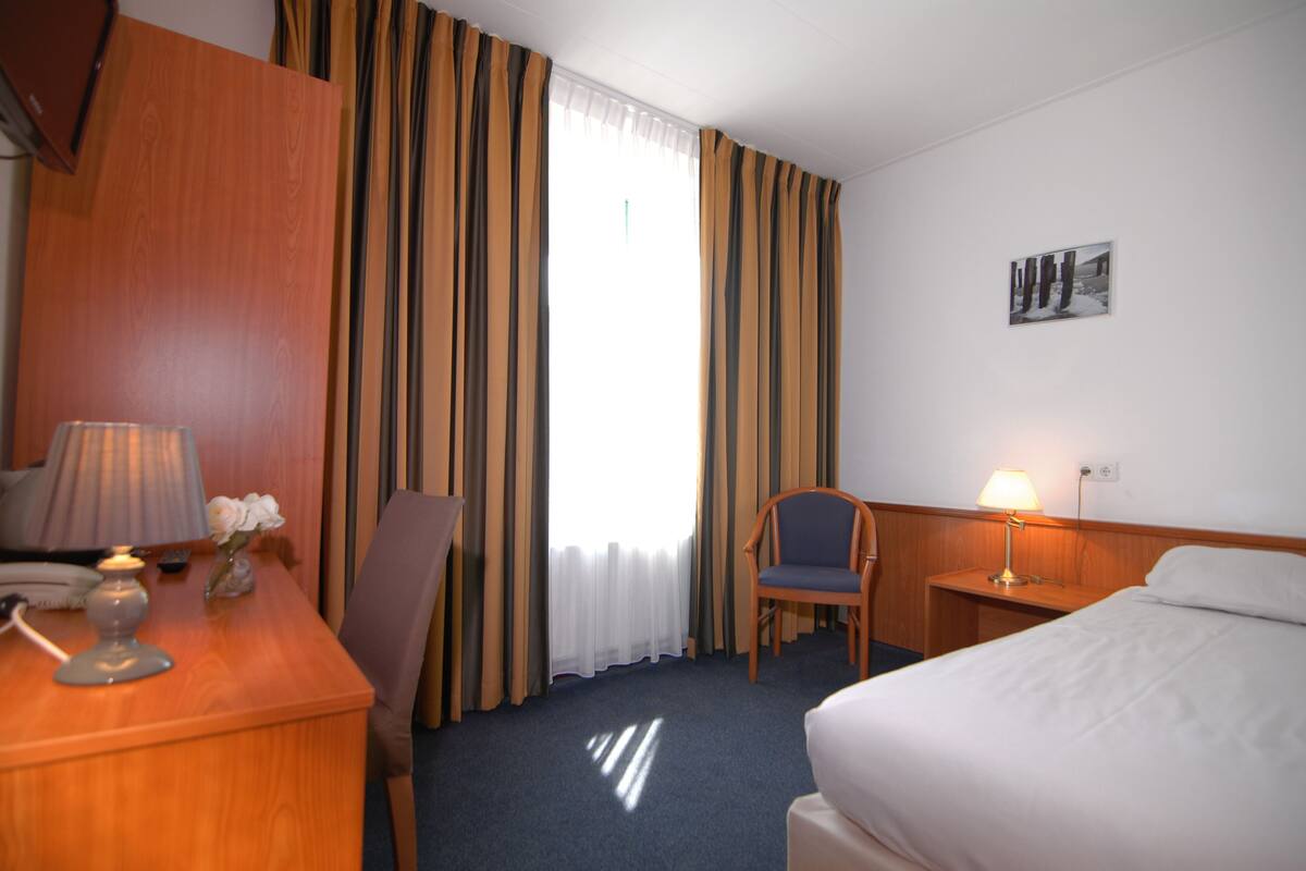 Ett hotellrum med ett skrivbord i trä, en stol, en säng, ett nattduksbord och en lampa.